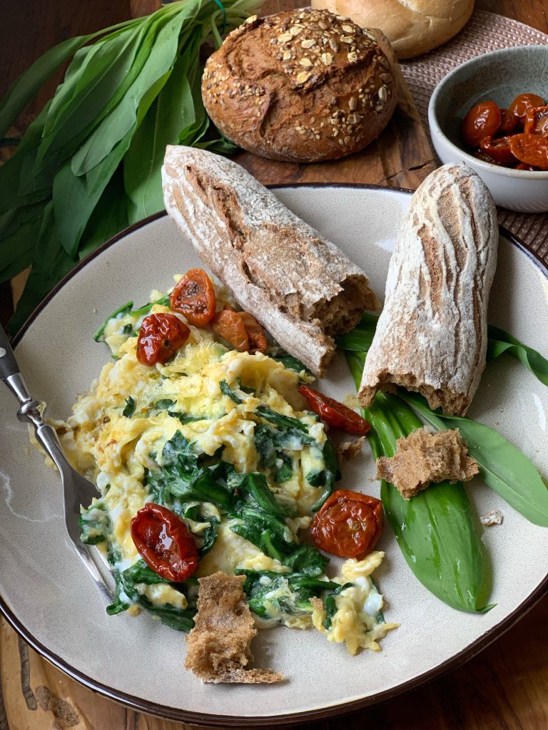 Rührei mit Bärlauch, Frühlingszwiebeln, eingelegten Tomaten und Brot auf einem Teller. 