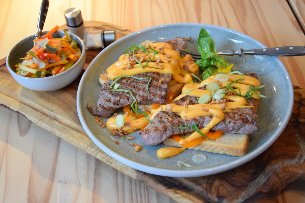 Toastscheiben mit Roastbeef, Röstzwiebeln und Sauce belegt auf einem Grauen Teller. Daneben steht eine kleine Schüssel mit eingelgtem Gemüse. Salz und Pfeffer stehen daneben.