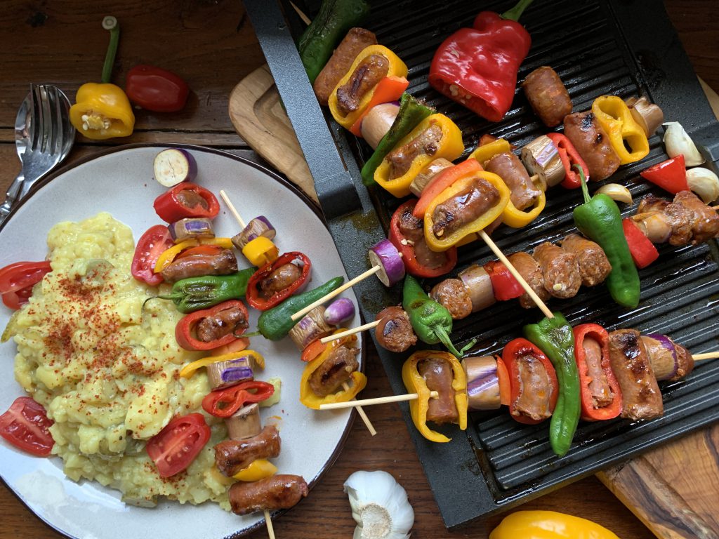 Merguez-Gemüsespieße und Kartoffelsalat auf einem Teller. Daneben ist eine Grillpfanne mit weiteren Spießen. Das Bild ist von oben fotografiert.