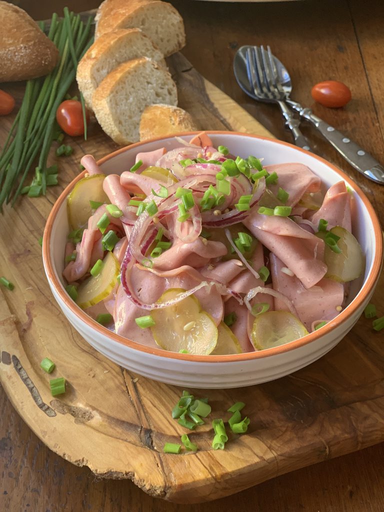 Wurstsalat mit Essiggurkenscheiben und Zwiebeln in einer Schüssel. Daneben sind Schnittlauch, Brotscheiben und Besteck zu sehen. Ein paar Mini-Tomaten sind ebenfalls auf dem Tisch.