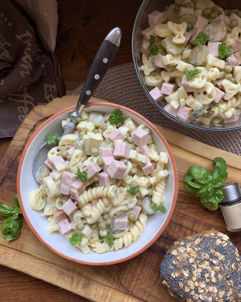 Eine kleine Schüssel Nudelsalat auf einem Holzbrett, der auf einem Tisch steht. Ein Löffel steckt in der Schüssel. Daneben steht die große Schüssel mit Nudelsalat. Am rechten unteren Bildrand sieht man einen Salzstreuer, ein paar Basilikumblätter und ein Stück Brötchen.