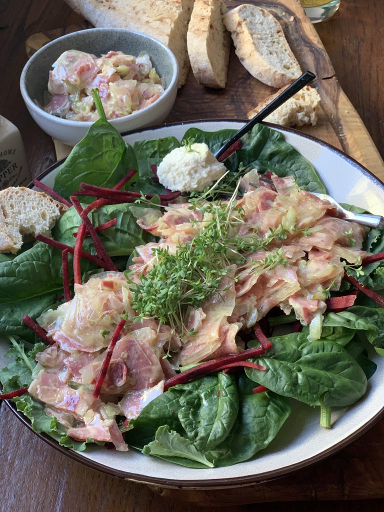 Vespersalat mit Schweinebäckle & Zwiebeln auf einem Salatbett auf einem Teller serviert. Daneben ist ein zusätzliches Schälchen Schweismaulsalat sowie ein Weißbrot und zwei abgeschnittene Scheiben. Am Teller ist ein Löffel mit Sahnemeerrettich zu sehen.