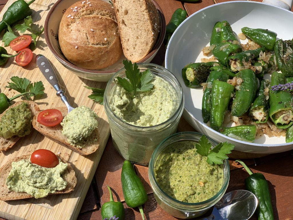 Auf einer Holzplatte sind Brotscheiben mit Dipps bestrichen. Die beiden Dipps und ein und halb Brötchen in einer Schüssel sind neben dem Brett. Eine Pfanne mit Bratpeperonis steht ebenfalls auf dem Tisch.
