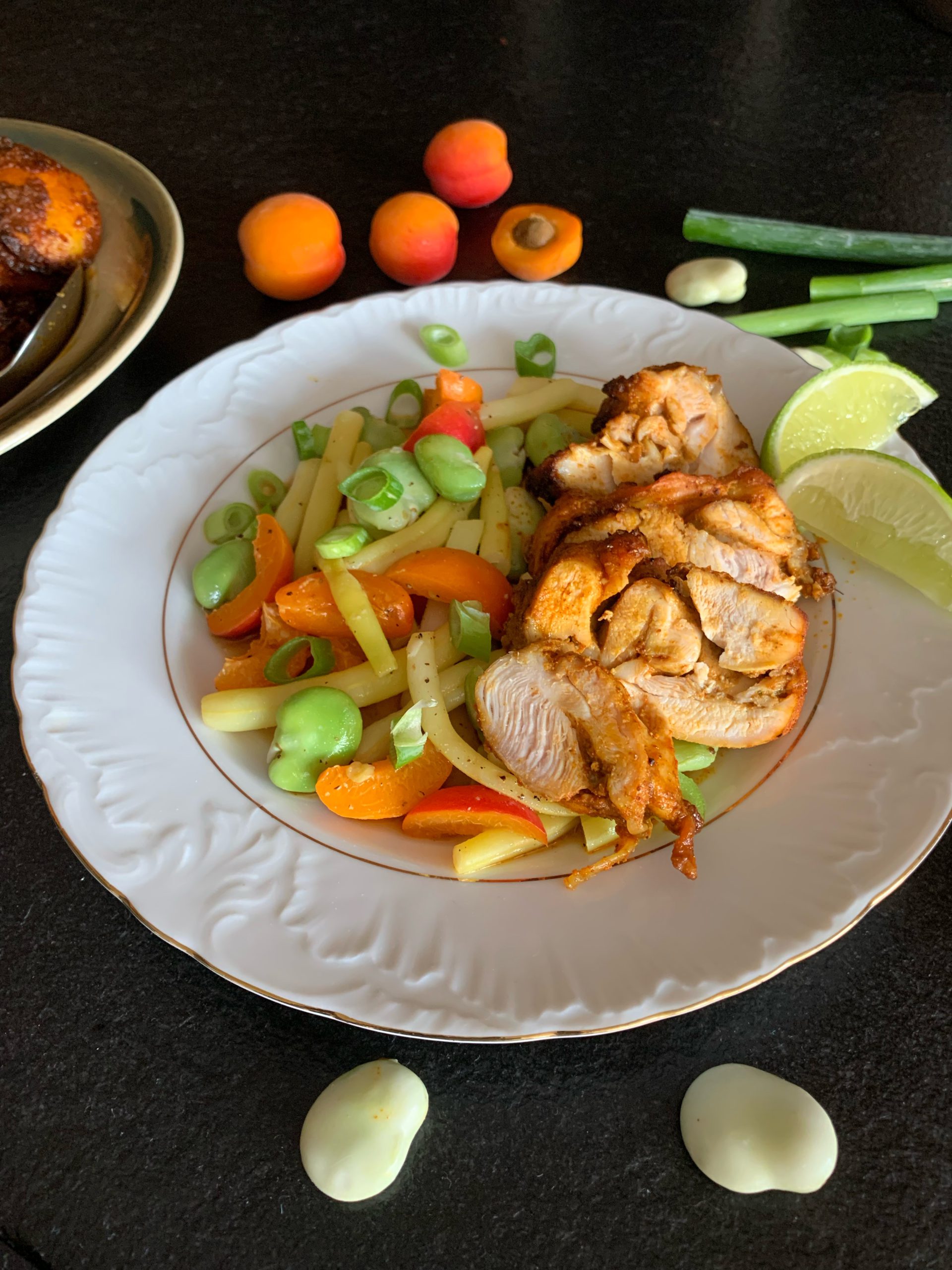 Pollo fino mit Bohnensalat mit Aprikosen auf einem Teller, der auf einem schwarzen Untergrund serviert wird. Daneben sieht man 4 Aprikosen und eine zerteilte Frühlingszwiebel.