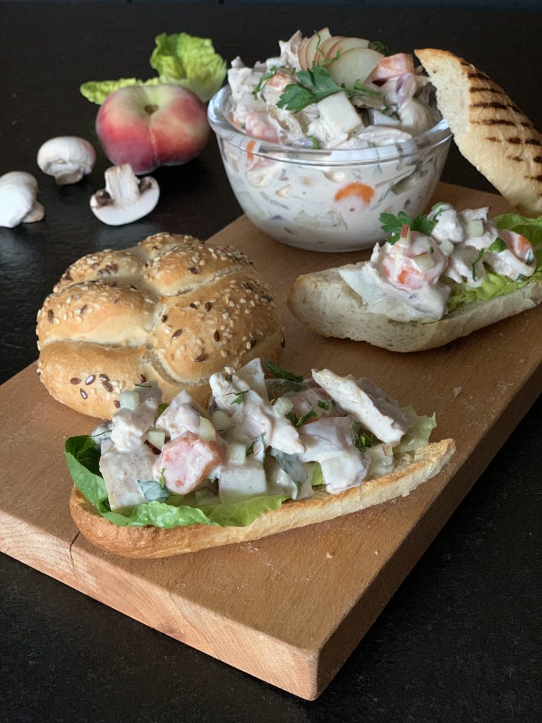 Auf einem Brett sieht man zwei Brotscheiben mit Geflügelsalat, eine Schüssel mit Geflügelsalat und ein Brötchen. Das Brett steht auf einem dunklen Untergrund. Neben dem Brett sieht man ein drei Champignons, einen Pfirsich und ein paar Salatblätter im Hintergrund.