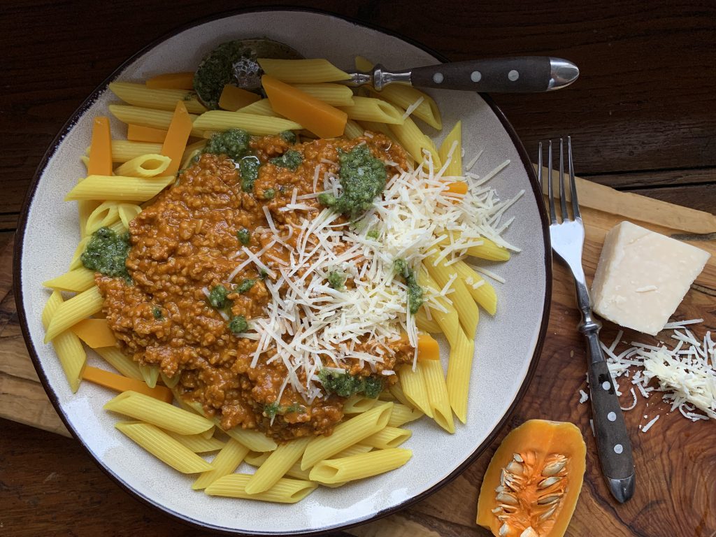 Curry-Nudeln mit Kürbisbolognese auf einem Teller. Ein Löffel liegt auf dem Teller und daneben liegt eine Gabel. Ein stück Kürbis und ein Stück Käse liegen neben der Gabel