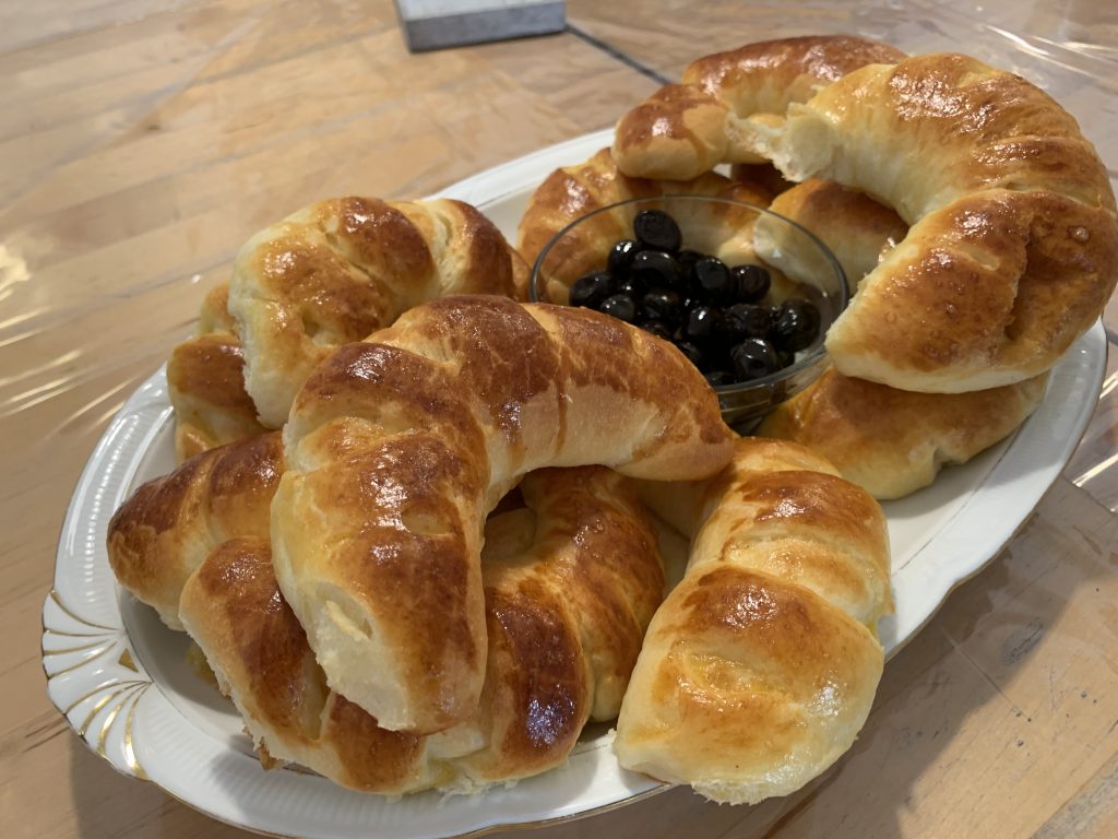 Zehn Gebäckteilchen in Mondform, gefüllt mit Käse auf einem weißen ovalen Teller. In der Mitte ist eine kleine Glasschüssel mit schwarzen Oliven. Der Teller steht auf einem braunen Tisch.