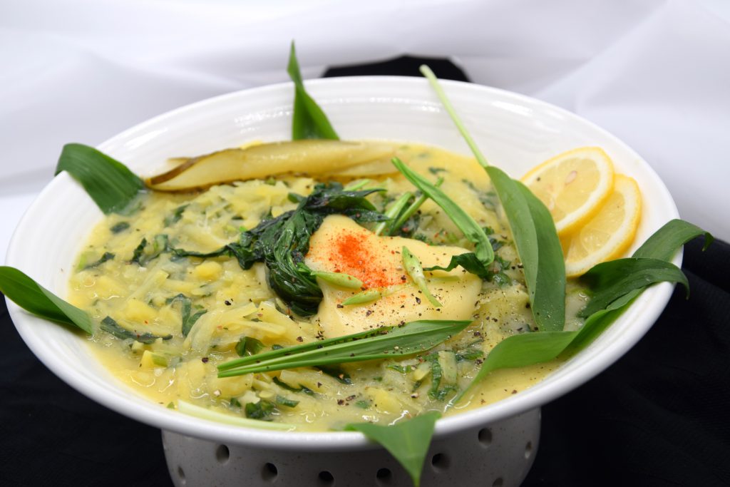 Kartoffelraspel – Risotto mit Käse und Bärlauch in einer Servierschale mit einem Sockel. Dekoriert wurde das Gericht mit geschnittenem Bärlauchblättern, Käsewürfeln und Zitronenscheiben. 