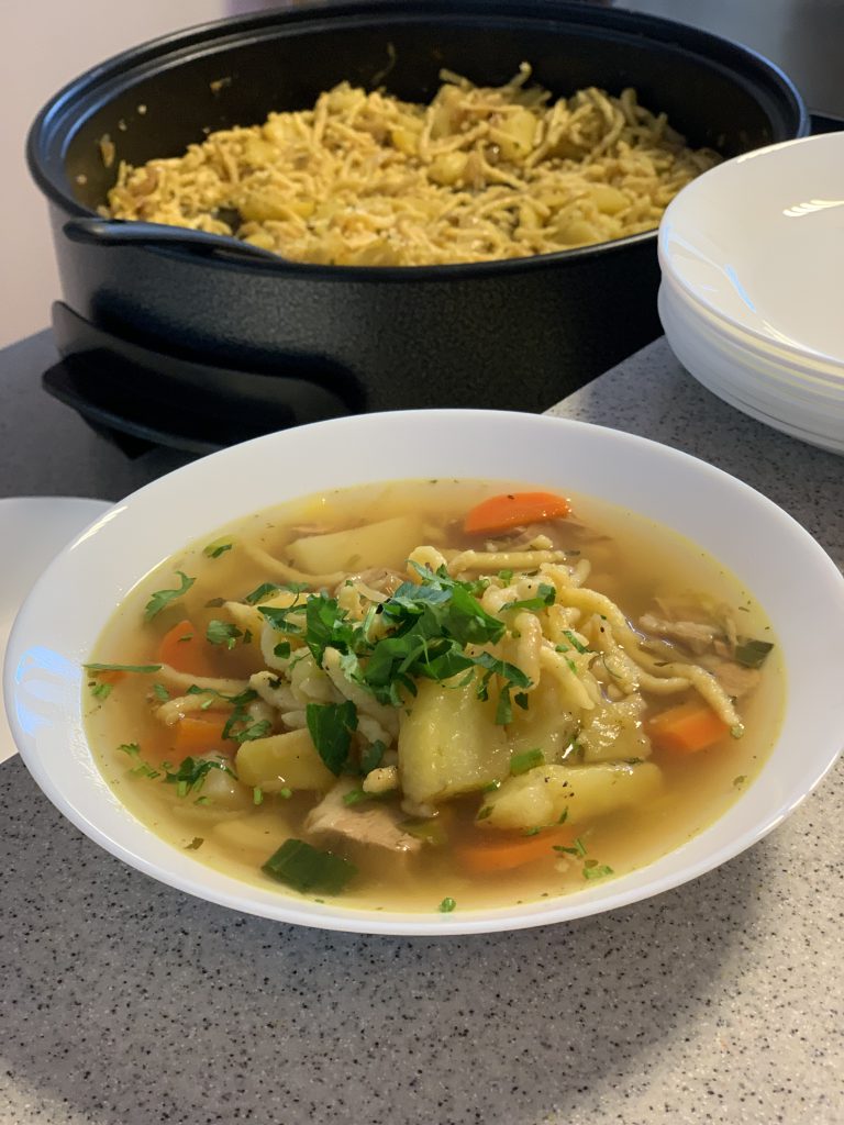 Auf dem Bild ist ein tiefer Suppenteller mit dem Eintopf Gaisburger Marsch zu sehen. Es schwimmen Spätzle, Karottenstücke, Kartoffelstücke und Fleischstücke in einer hellen Brühe. Garniert mit schnittener Petersilie. Der Teller steht auf einem grauen Untergrund. Hinter ihm steht ein großer schwarzer Topf mit dem restlichen Eintopf. Im oberen rechten Bildrand sind weitere Suppenteller, gestapelt zu erkennen.