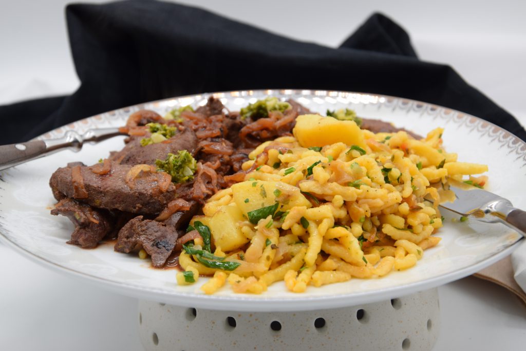 Auf einem Teller liegen auf der linken Seite gebratene Leberstücke in Zwiebelsoße und auf der rechten Seite sind Spätzle mit Kartoffelstücken gemischt. Das Gericht Kräutern bestreut. Links und rechts sieht man Messer und Gabel am Teller angelehnt. Hinter dem Teller ist eine schwarze Stoffserviette. Der Teller steht auf einem gepunkteten Sockel.