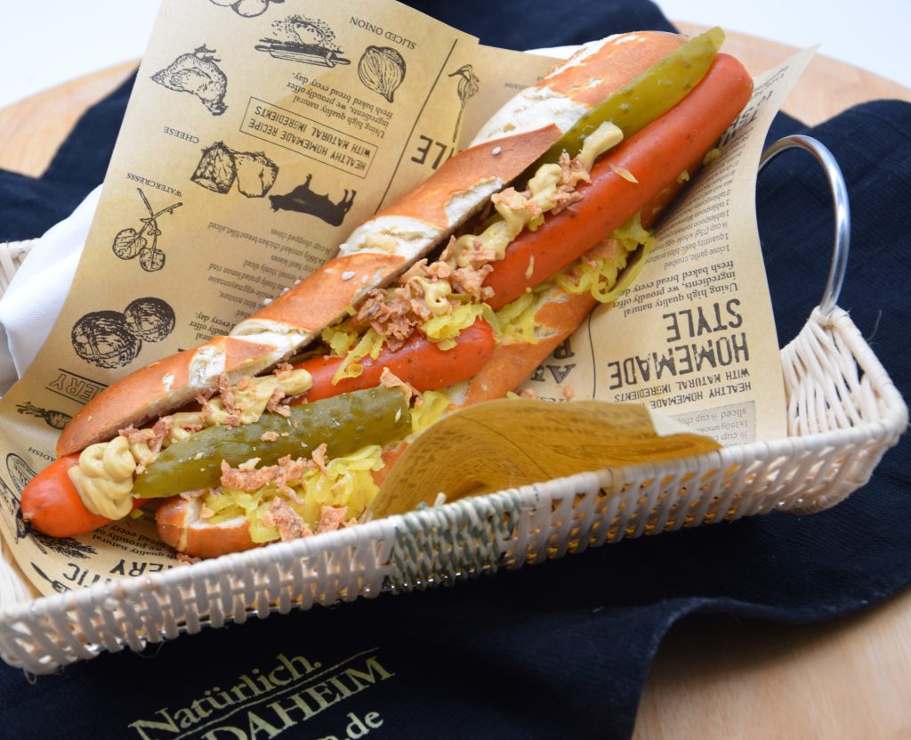 Längs aufgeschnittene Laugenstange in der zwei kleine Debrezinerwürstchen und Saure Gurkenstücke stecken. Mit Röstzwiebeln und Senf gewürzt. Der Hot Dog liegt auf einem Metzgerpapier in einem weißen Körbchen, das auf einem schwarzen Geschirrtuch steht. Auf dem Tuch steht in goldener Schrift Natürlich. Von Daheim. 