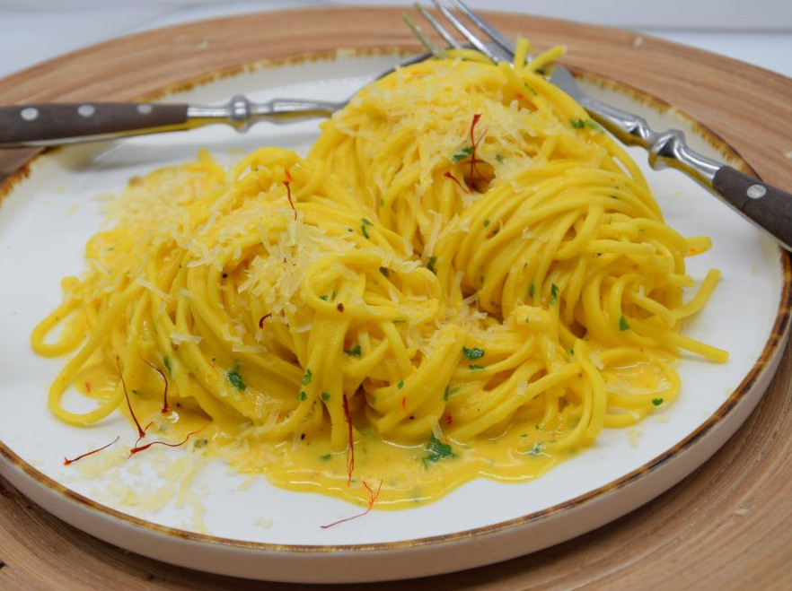 Spaghettinudeln in Safrankäsesoße auf einem flachen Teller. Man sieht Safranfäden auf den Nudeln. Der Teller steht auf einem hellbraunem Platzdeckchen. Am Tellerand sind Löffel und Gabel.