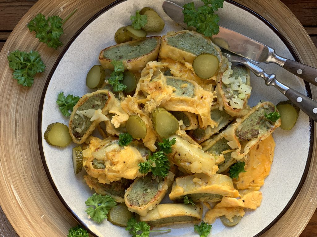 In Streifen geschnittene Maultaschen, die mit verquirlten Eiern gebraten wurden, auf einem Teller. Mit Essiggurkenscheiben und geschnittener Petersilie garniert. Messer und Gabel sind am Tellerrand angelehnt.