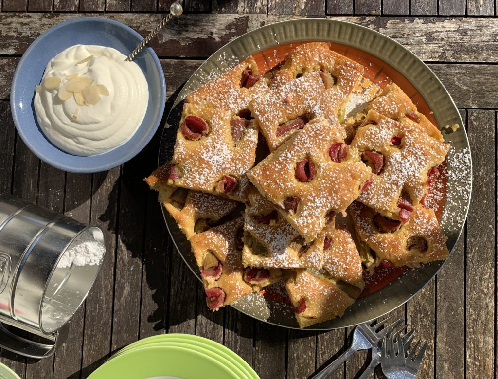 Rhabarberkuchen auf einem Servierteller. Links daneben ist eine kleine Schüssel mit Sahne. AM unteren Bildrand kann man grüne Teller und drei Gabeln erkennen.