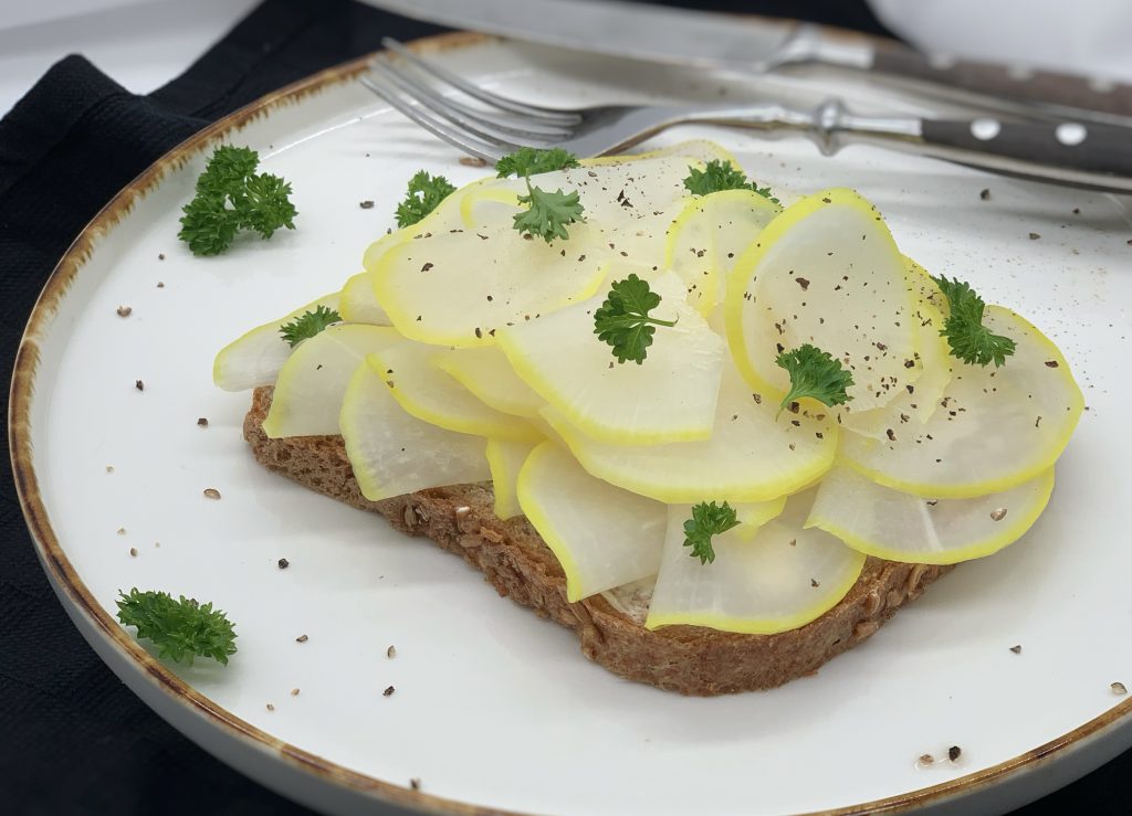 Eine Scheibe Volkornbrot mit Mairübchenscheiben belegt. Darüber ist etwas geschnittene Petersilie gestreut. Das Brot liegt auf einem weißen Teller. Am Tellerrand liegen Messer und Gabel. Der Teller liegt auf einem schwarzen Untergrund.