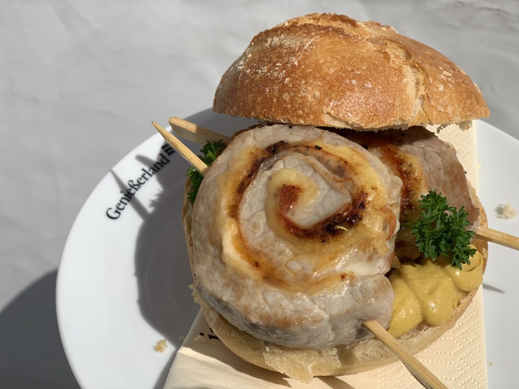 Ein Brötchen gefüllt mit zwei Schweinefiletrouladenscheiben, die auf kleinen Spießchen stekcen. Garniert mit Senf und Petersilie. Das Brötchen liegt auf einem weißen Teller mit dem kleinen schwarzen Logo"Genießerland"