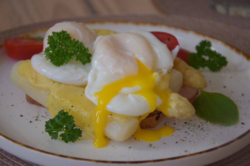 Toastbrotscheibe mit Spargelstücken, Schinken, Käse und pochiertem Ei belegt. Der Toast liegt auf einem runden, weißen Teller mit niedrigem Rand und ist mit Petersilie garniert.