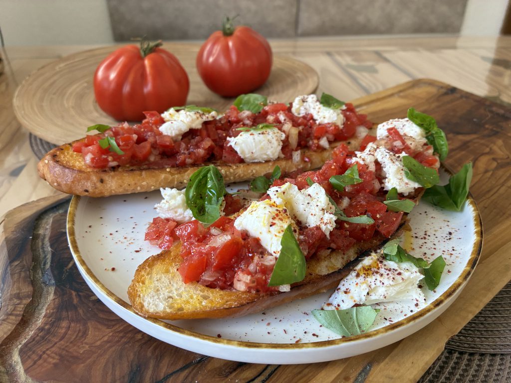 Zwei Scheiben Röstbrot belegt mit klein geschnittenen Tomaten, Mozzarella und gezupften Basilikumblättern auf einem runden Teller. Der Teller steht auf einem Holzbrett. Im Hintergrund sieht man zwei Oschenherztomaten in einer großen, falchen Holzschale.