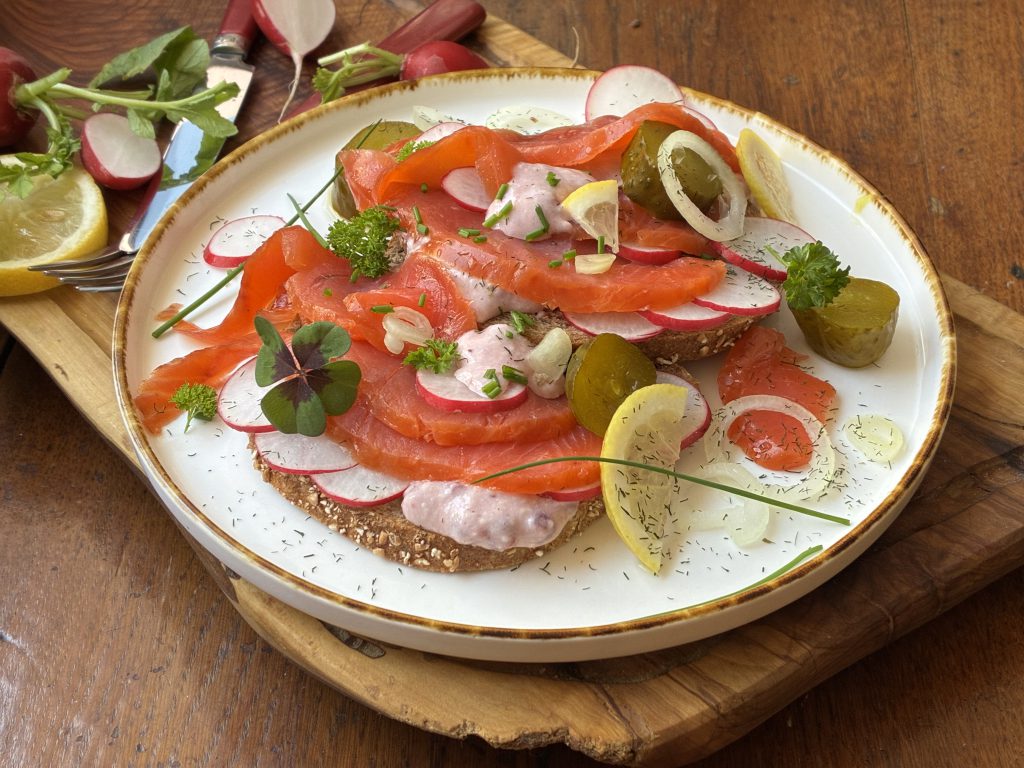 Brotscheibe belegt mit gerächerten Lachsforellenstücken, Preiselbeer-Meerrettich, Radieschenscheiben und Gurkenscheibchen. Garniert mit Petersilie, Zwiebelringen und Zitronenvierteln. Der Teller steht auf einem Holzbrett. Auf dem Holzbrett sieht man ein Messer und Radieschen.
