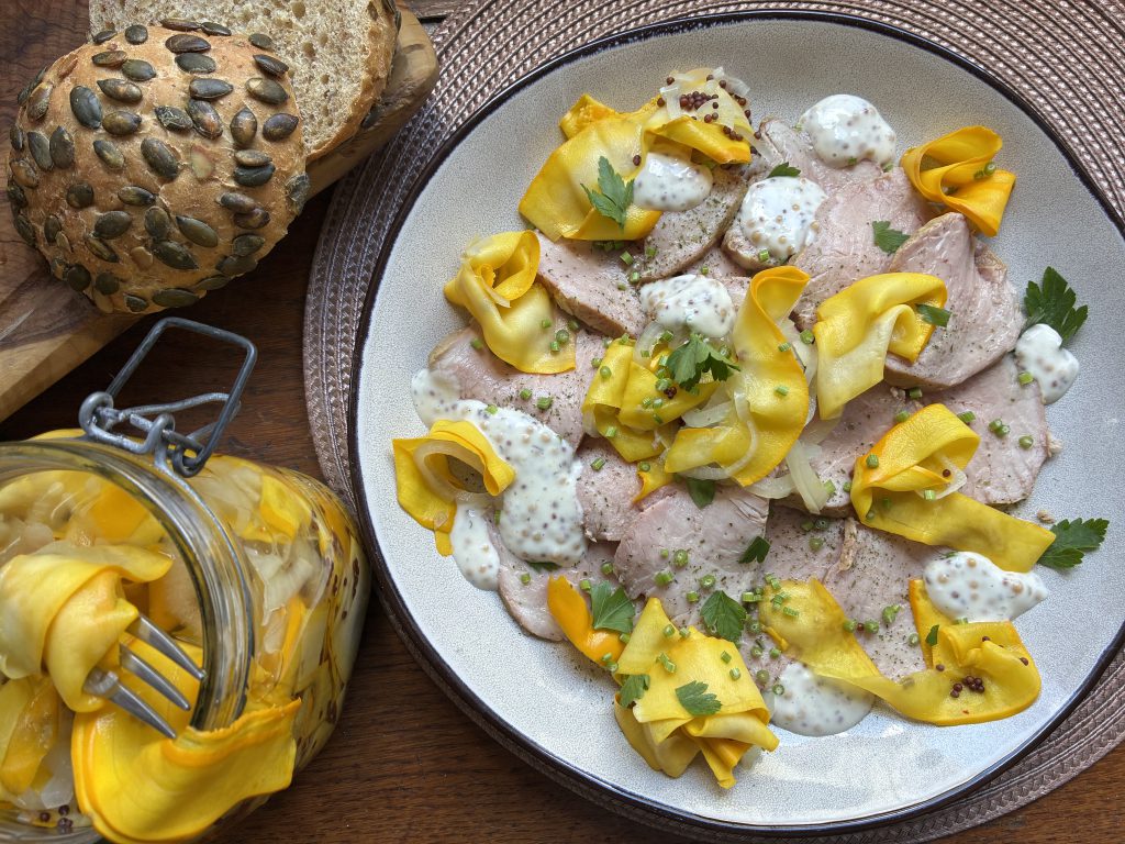Kalbsrückenscheiben mit eingelegten, gelben Zucchinistreifen auf einem flachen Teller. Mit Ganzkorn-Mayo und geschnittener Petersilie garniert. Neben dem Teller liegt ein aufgeschnittenes Kürbiskernbrötchen. Das Einmachglas mit Zucchini ist ebenfalls auf dem Bild.