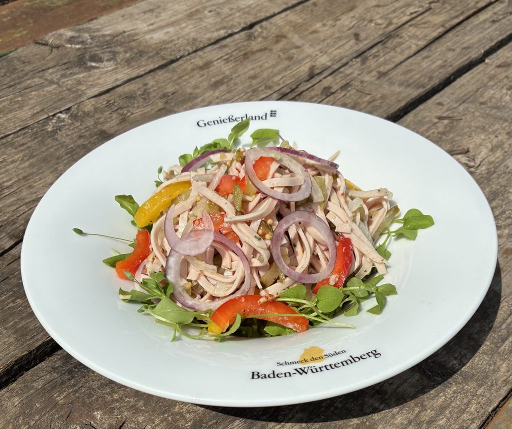 Fleischsalat in einem tiefen Teller mit Aufschrift "Gnießerland" und dem SdS-Logo. Der Salat ist mit Zwiebelringen, Paprikastücken und Kresse.