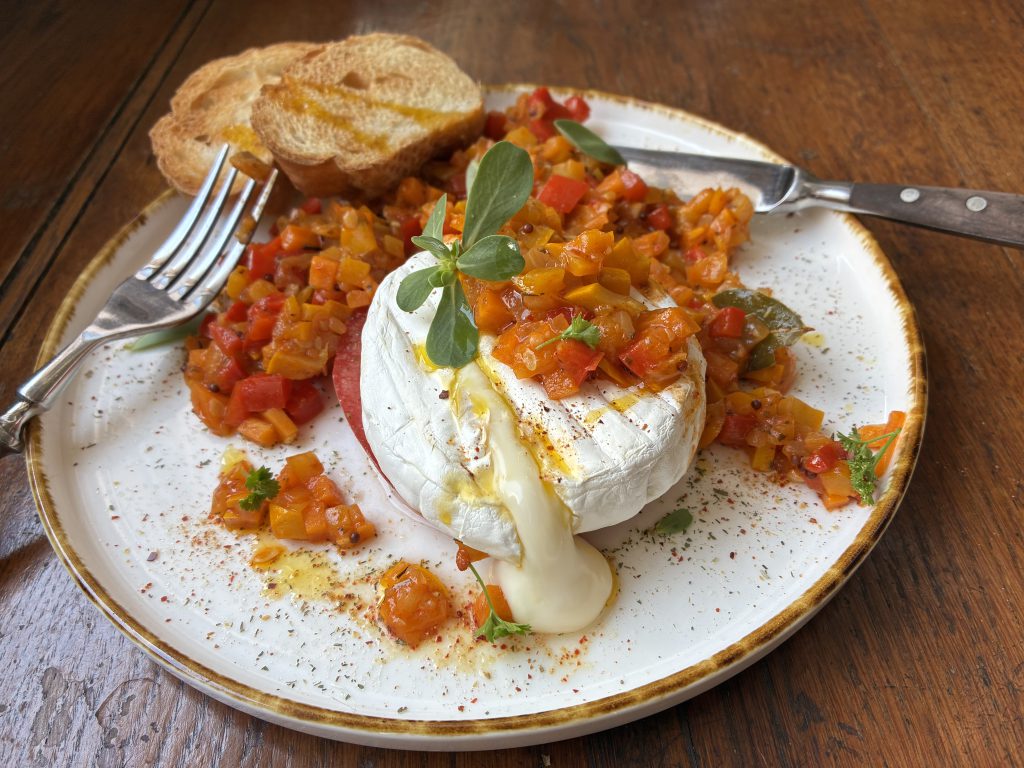 Gebratener Camembert mit Tomate und Ratatouille auf einem weißen Teller. Rechts am Tellerrand liegt eine Gabel, rechts ein Messer. Auf dem Teller sind ebenfalls zwei Scheiben eines Baguettes.
