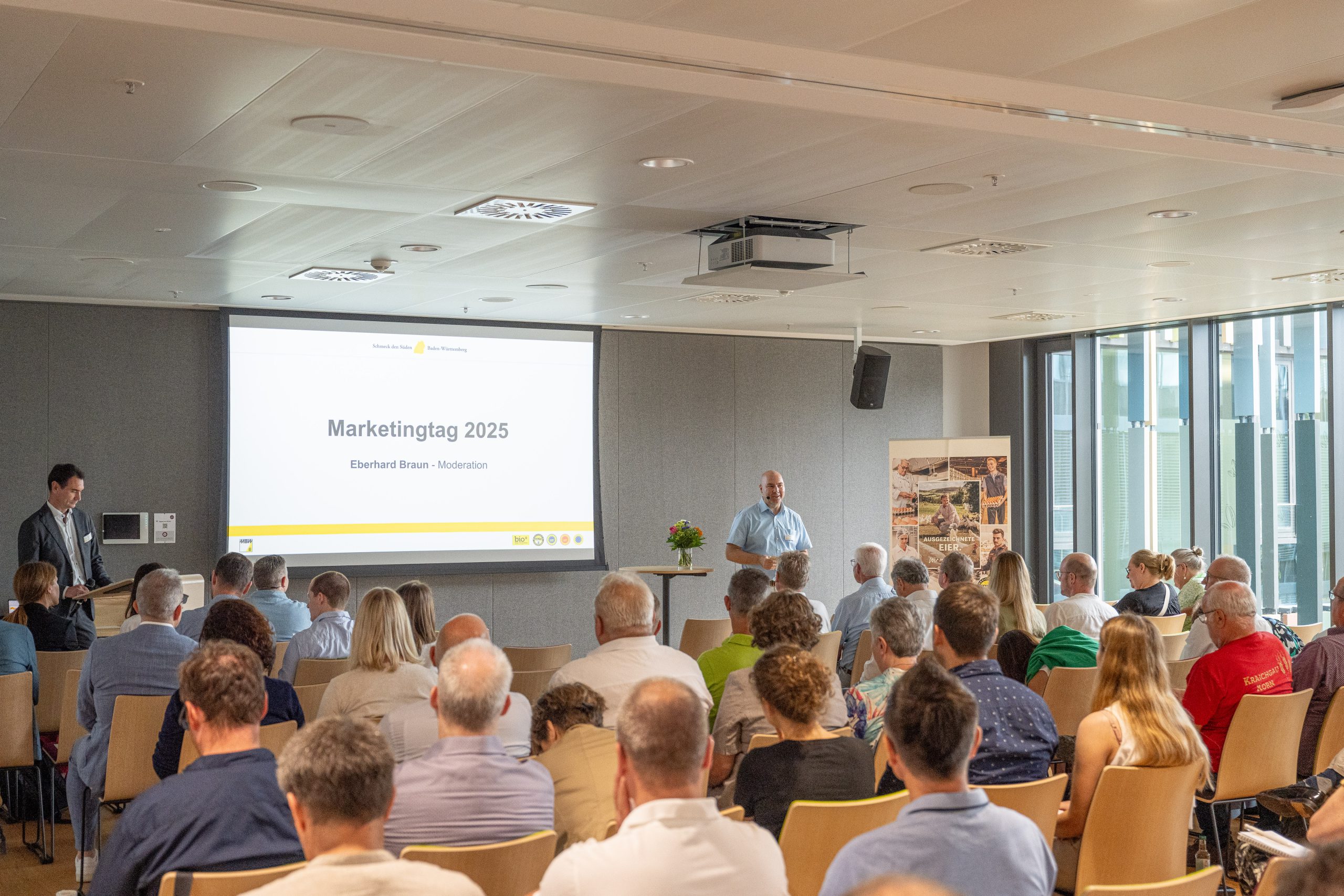Veranstaltungsraum mit Leinwand, auf der Marketingstag 2025 steht. Eberhard Braun ist ebenfalls vor dem versammeltem Publimkum