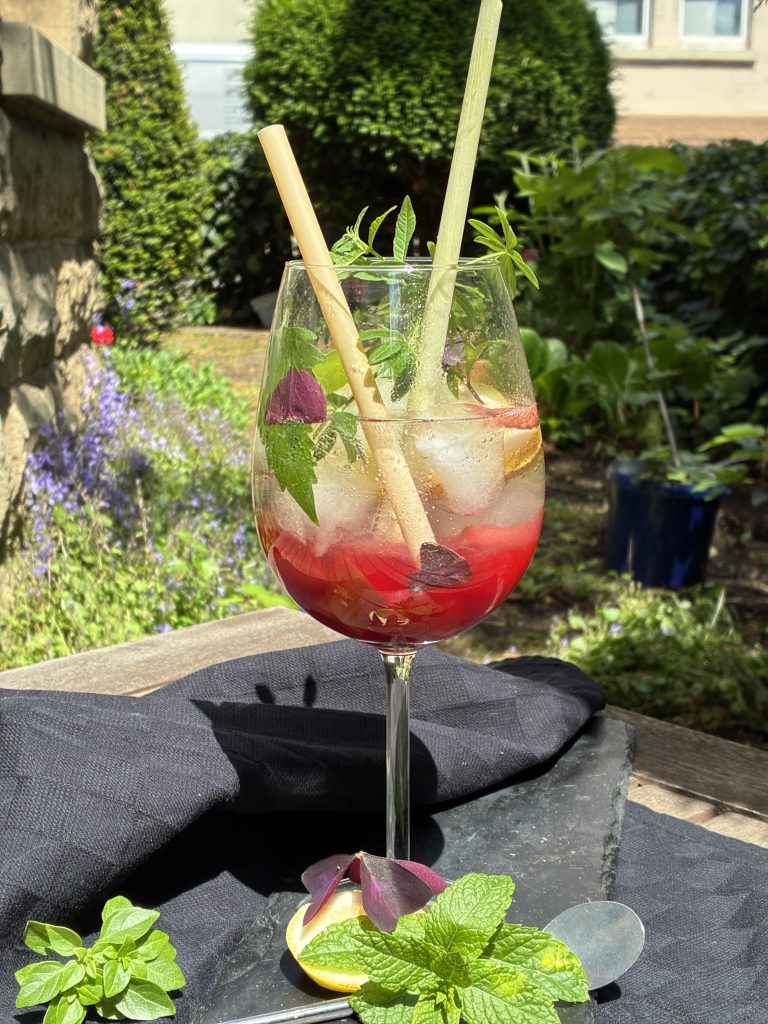 Gin-Cocktail mit roten Beeren, Kräutern und Eiswürfeln in einem Weinglas. Im Weinglas stecken zwei Strohhalme. Vor dem Glas sind kleine Büschel von Basilikum und Minze sowie ein Cocktaillöffel. Das Glas steht auf einer schwarzen Tischdecke auf einem Holztisch in einem Garten. Im Hintergrund sind lilafarbene Blumen sowie verschiedene Büsche.
