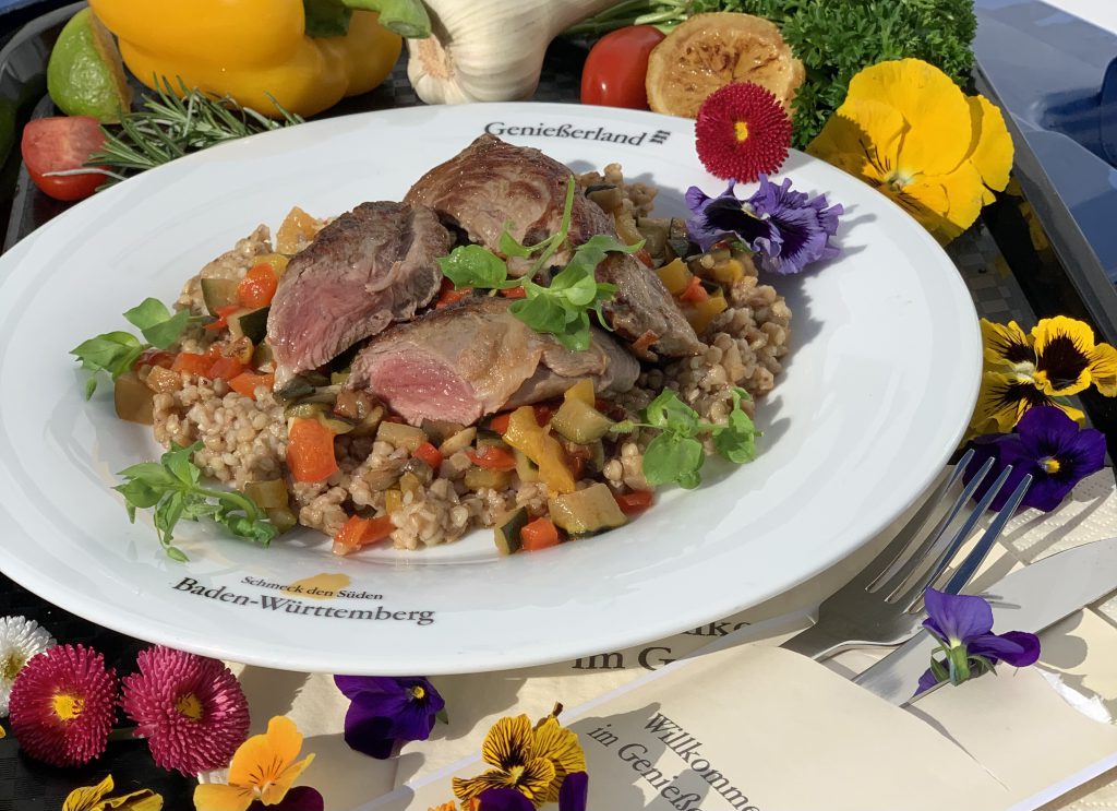 Gebratene Stücke von Württemberger Lammfilet auf Buchweizen-Risotto und Ratatouillegemüse auf einem weißen Teller mit dem SdS-Logo und der Aufschrift Genießerland. Der Teller ist garniert mit verschiedenen bunten Blüten, auch außerhalb des Tellers. 