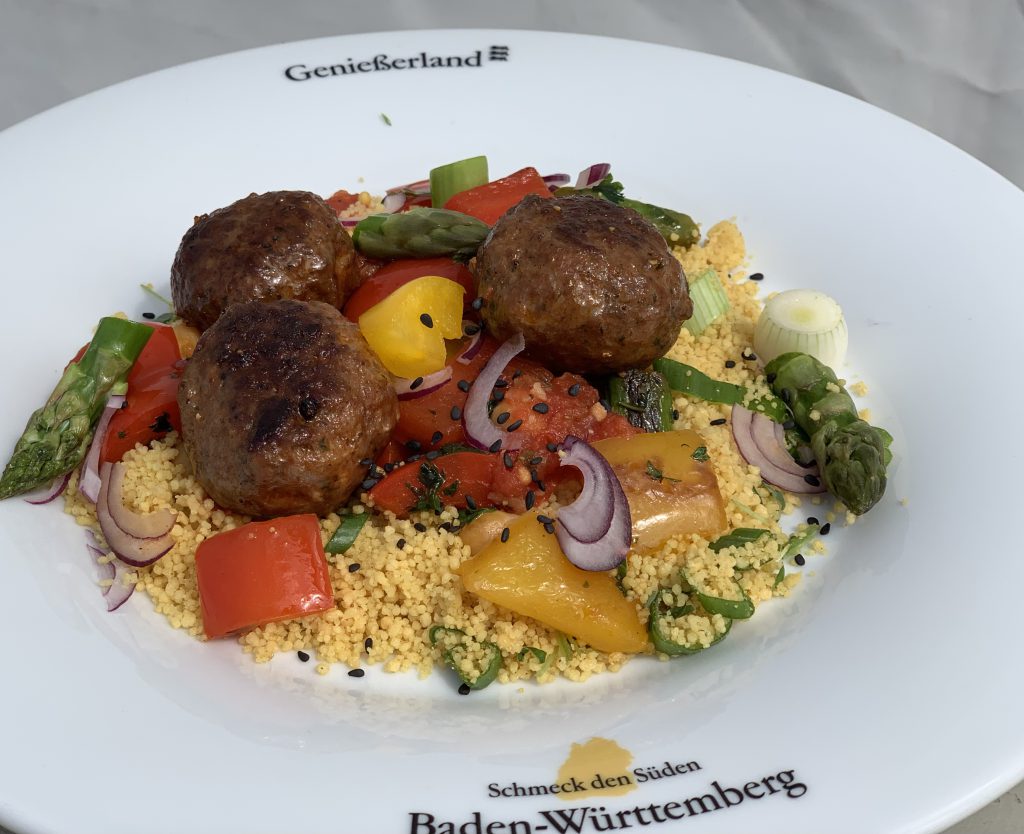 Drei kleine Frikadellen auf verschiedenem Gemüse und Couscous. Garniert ist das Gericht mit frischer Minze und Petersilie Der Teller ist weiß mit der Aufschrift Genießerland und dem "Schmeck den Süden"-Logo.