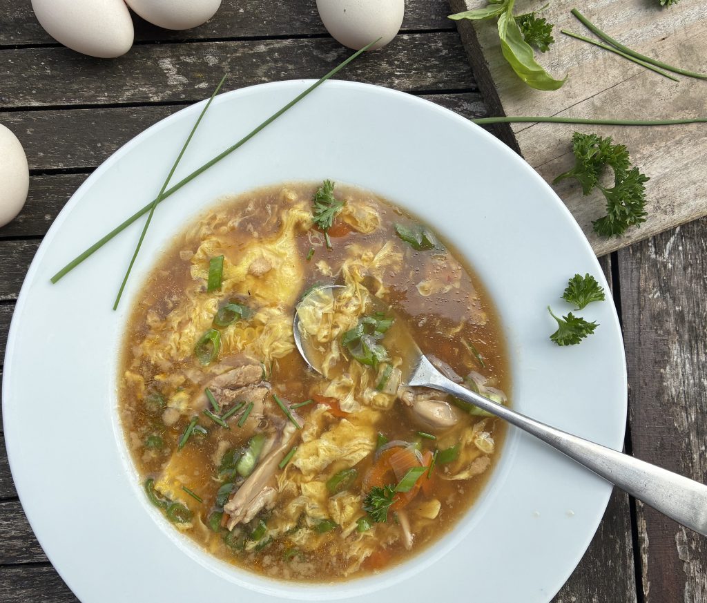 Ein tierfer Teller mit Hühnerbrühe, Hühnchenfleisch, Gemüse und Eierstich. Im Teller liegt seitlich ein Löffel in der Suppe. Die Suppe ist garniert mit Schnittlauch und Petersilie. Auch am Tellerrand sind zwei Schnittlauchhalme und etwas geschnittene Petersilie. Der Teller steht auf einem grauen Holztisch. Daneben sind vier Eier und eine Schneidebrett mit verschiedenen Kräutern am oberen Bildrand.