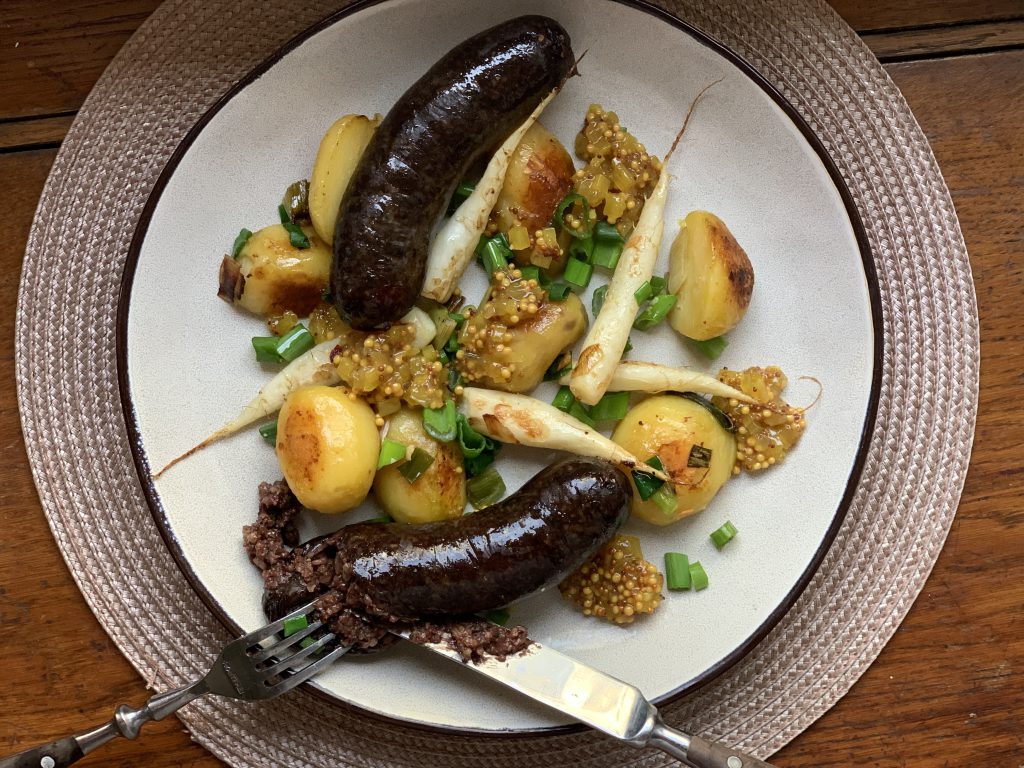 Zwei gebratene Schwarzwürste mit Rettich, Kartoffeln und Birnen-Senf-Chutney auf einem weißen flachen Teller. Garniert mit Frühlingszwiebelringen. Der Teller steht auf einem runden, beigen Platzdeckchen auf einem Holztisch. Das Bild wurde von oben fotografiert.