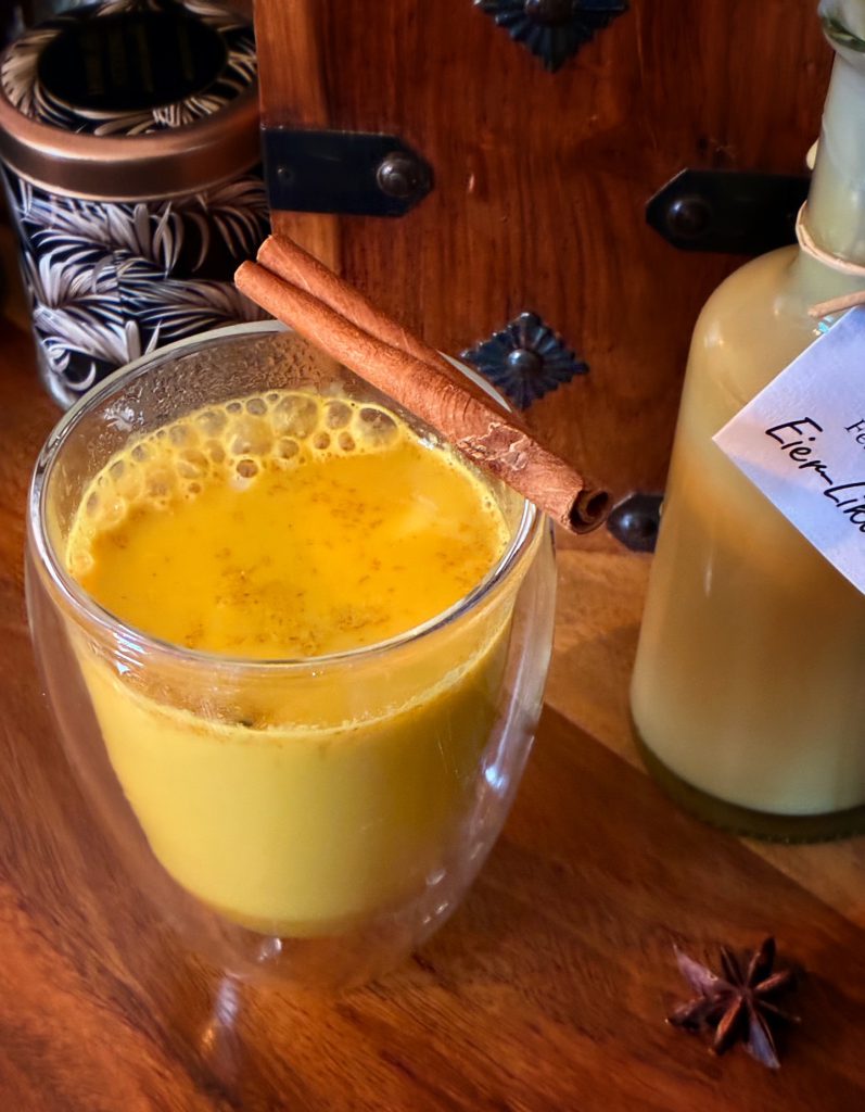 Goldene Milch in einer doppelwandigen Glastasse. Garniert mit einer Zimtstand am oberen Rand. Das Glas steht auf einem Holztisch und daneben ist eine angebrochene kleine Flasche mit Eierlikör und eine kleine Keksdose.