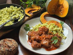 Kalbsgulasch und grüne Spätzlemit Kürbisspalten auf einem weißen Teler. Neben dem Teller sind auch ein angeschnittener Kürbis und Kürbisspalten, eine Pfanne mit Spätzle, Petersilie und ein Brötchen.