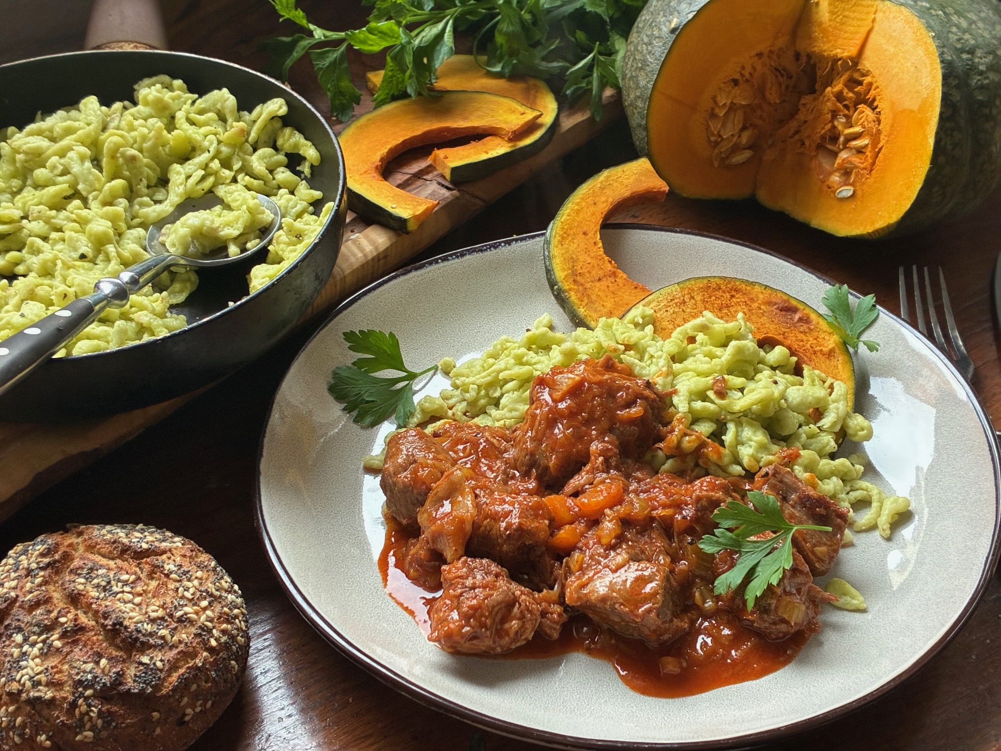 Kalbsgulasch und grüne Spätzlemit Kürbisspalten auf einem weißen Teler. Neben dem Teller sind auch ein angeschnittener Kürbis und Kürbisspalten, eine Pfanne mit Spätzle, Petersilie und ein Brötchen.