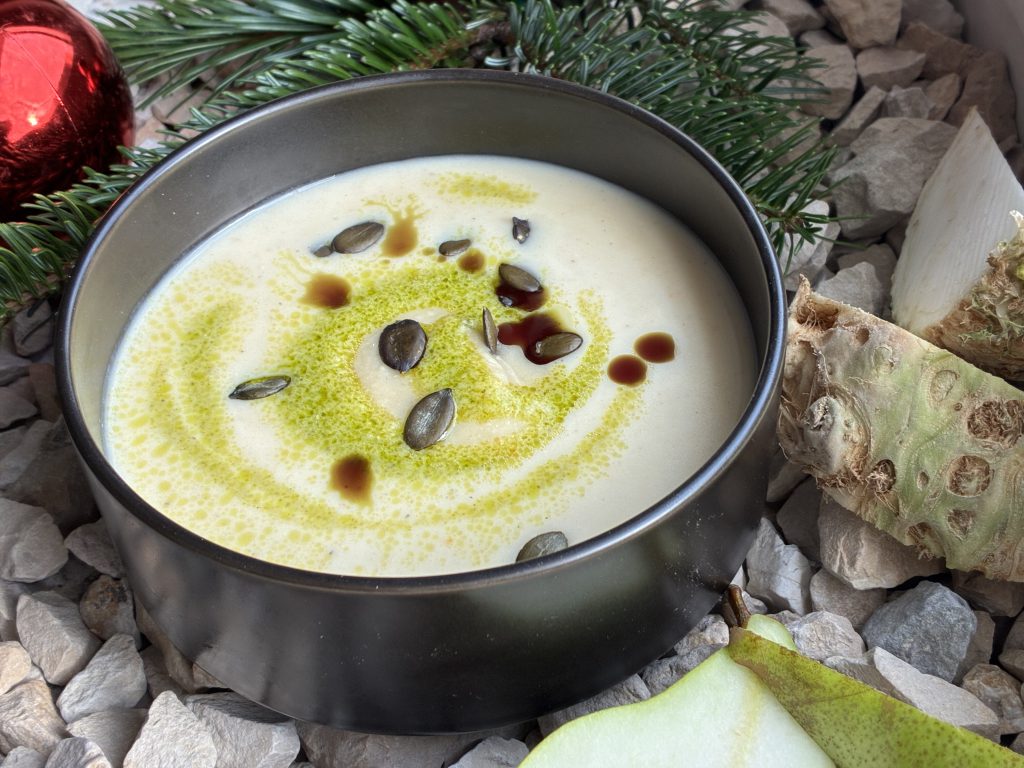 Pastinakencremesuppe in einem schwarzen Schüsselchen. Garniert mit Kürbiskernen und Kürbisöl. Das Schüsselchen steht dekorativ auf grauen Steinen. Daneben sind Selleriestücke und Birnenviertel abgebildet. Oben links sieht man eine rote Weihnachtskugel und einen Tannenzweig.